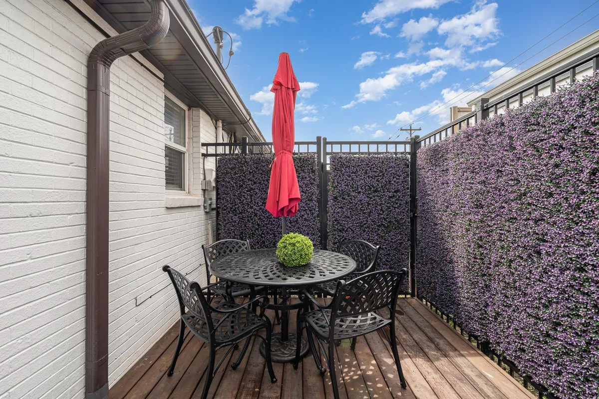 A cheerful patio scene featuring a round black table under a red umbrella, with a green plant centerpiece and a colorful floral wall in the background. The setting is ideal for outdoor dining or relaxed gatherings.