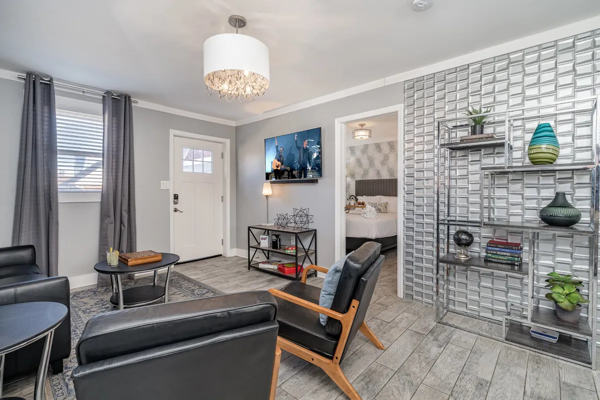 The image features a chic, light-filled living space with comfortable seating arranged around a stylish coffee table, a modern decorative shelving unit, an artistic light fixture overhead, and a large TV displaying art. The entrance door hints at a private, welcoming atmosphere, while the overall décor is warm and contemporary.