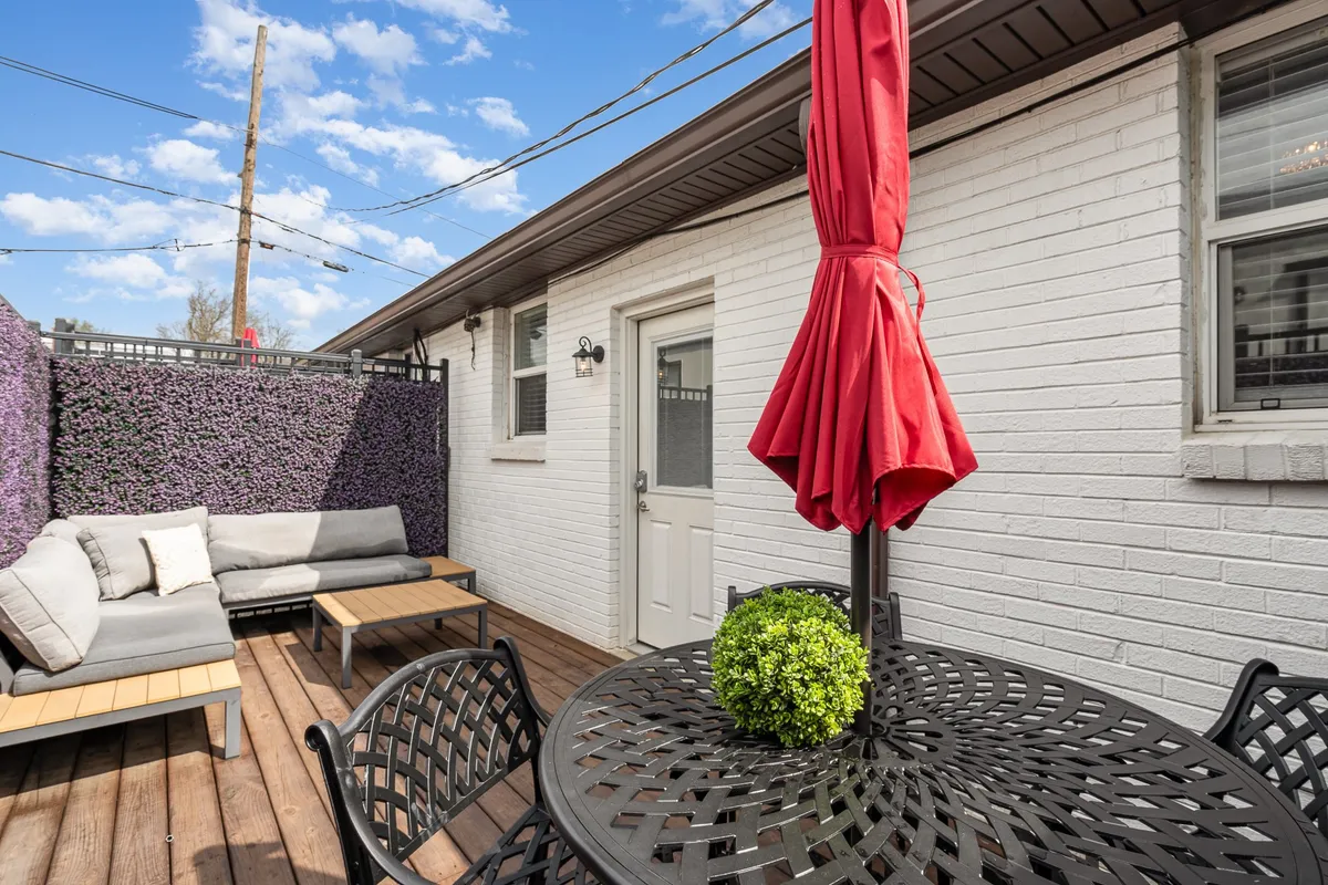 A cozy outdoor patio area featuring a red umbrella, green potted plants, and comfortable seating with a backdrop of a brick wall and greenery. The space is inviting and perfect for outdoor dining or relaxing together.