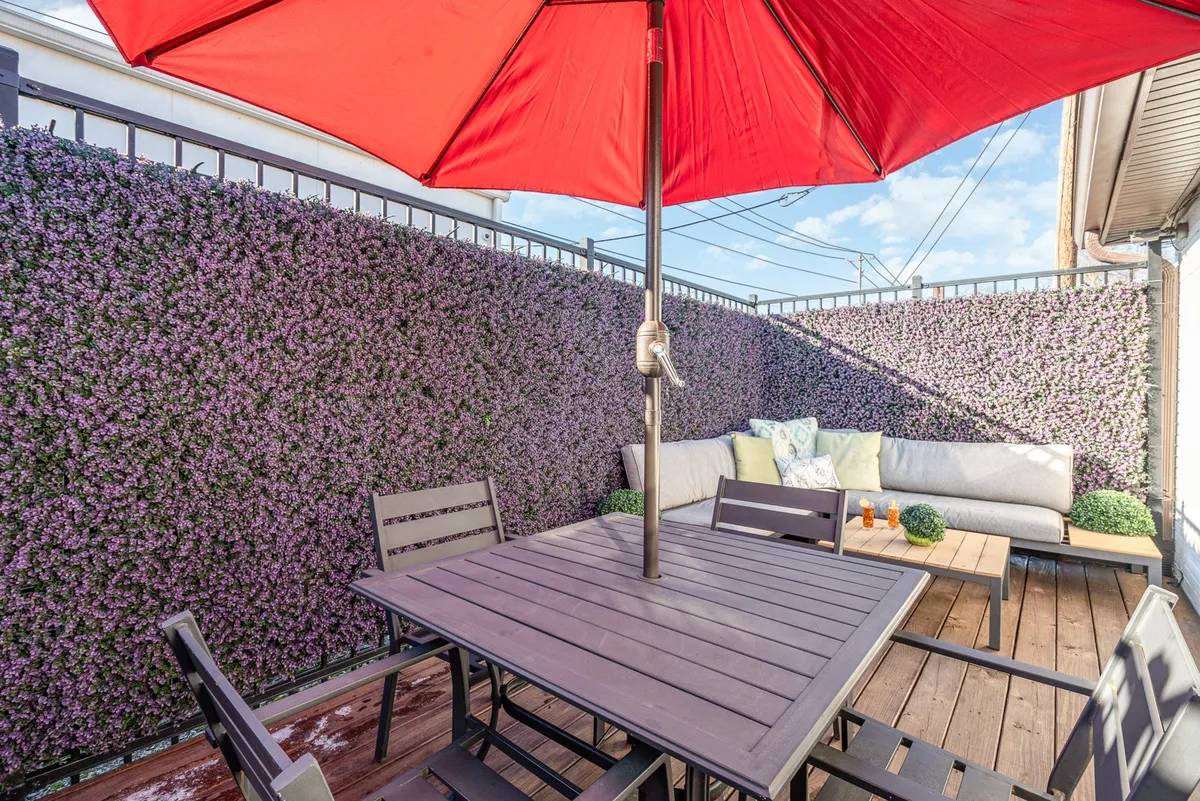 A cozy and intimate patio decorated with a red umbrella, green cushions on a wooden table, and a purple accent wall, inviting guests to relax, socialize, or enjoy a meal outdoors. The arrangement supports both small group gatherings and quiet personal moments.