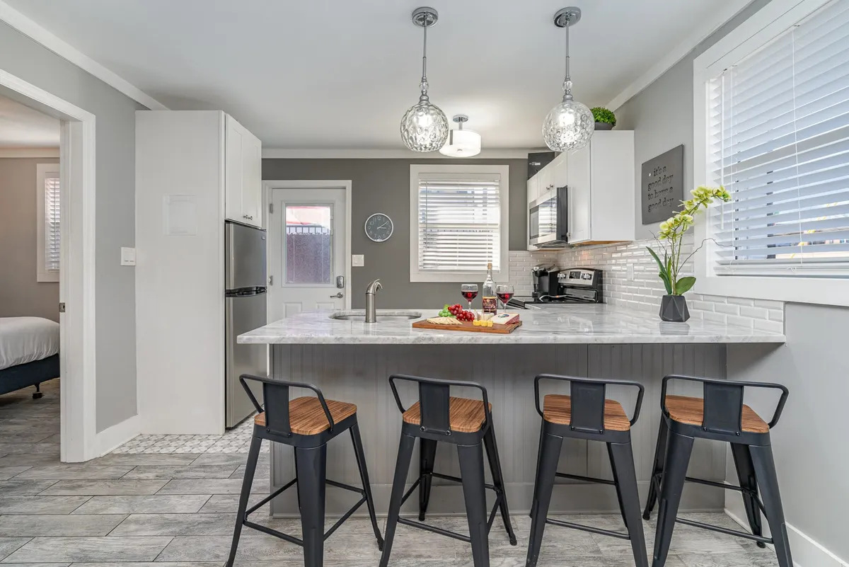 A contemporary kitchen featuring a kitchen island with bar stools, wine glasses ready for a toast, a fresh vegetable platter, and soft pendant lights. Stainless steel appliances are visible, creating a sense of a fully equipped, stylish space for entertaining.
