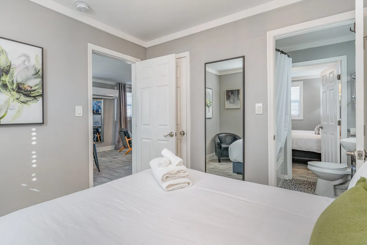 A tranquil bedroom with a neatly made bed, towels arranged on top, floral wall art, and a mirror that reflects the calm ambiance. The scene suggests a peaceful overnight stay with all essentials provided.