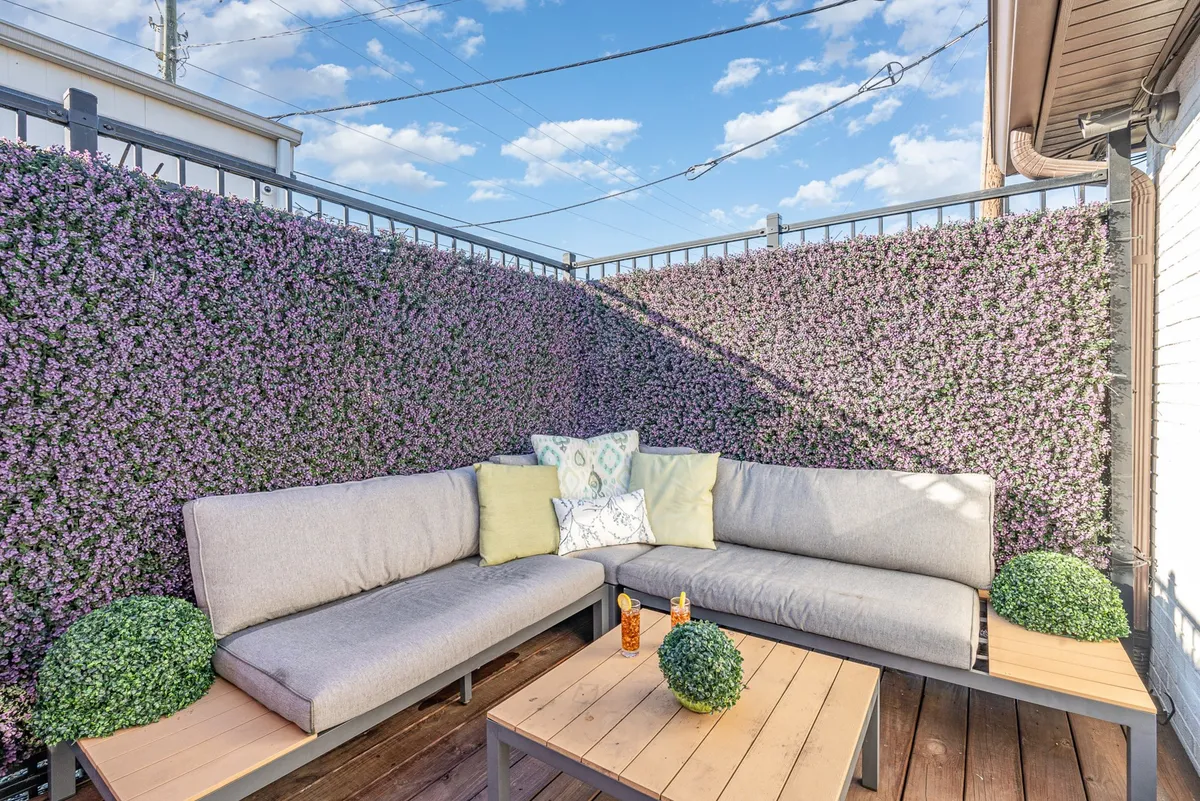 A tranquil outdoor seating area featuring a plush sofa with decorative cushions, a wooden coffee table, and touches of greenery. The patio is shaded and intimate, perfect for enjoying quiet mornings or evening conversations.