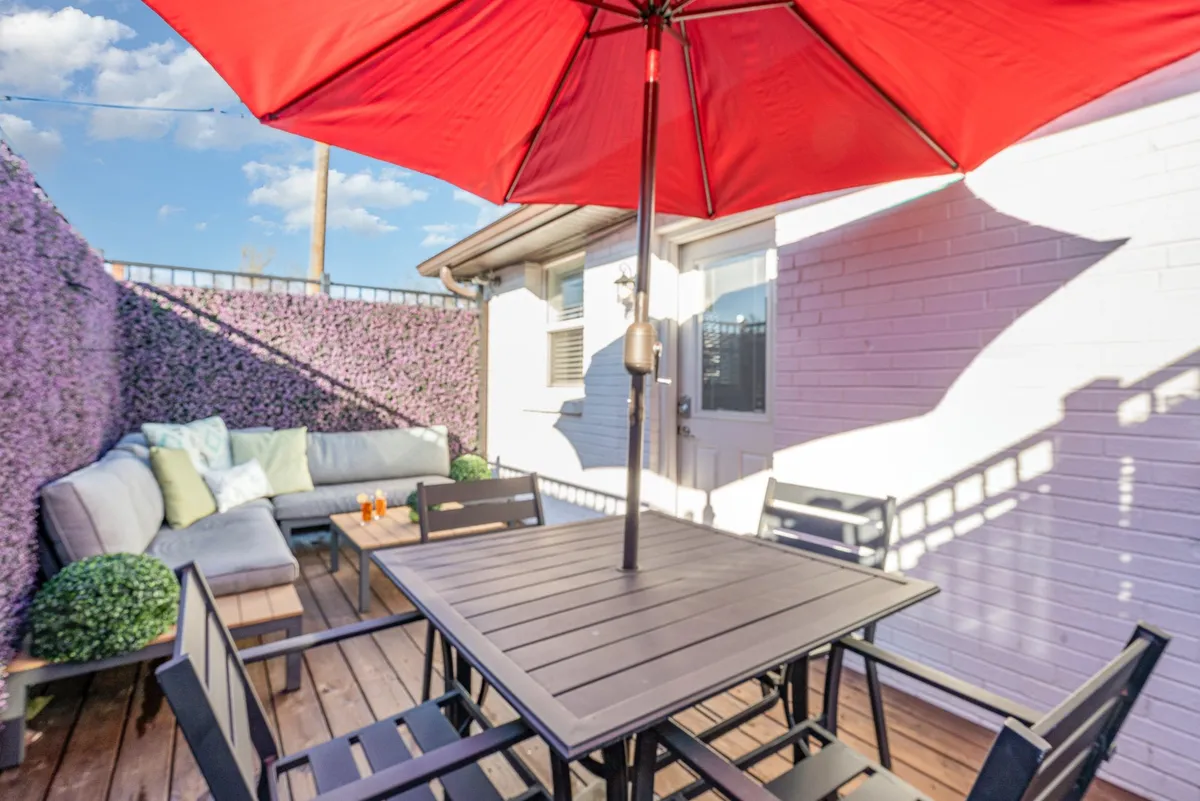 A cheerful outdoor patio with a vibrant red umbrella, a comfortable sofa seating area, and an outdoor dining table for four, all set up for relaxed dining and socializing. The setting includes accents like fairy lights and lush greenery, evoking an inviting atmosphere for evening or afternoon gatherings.