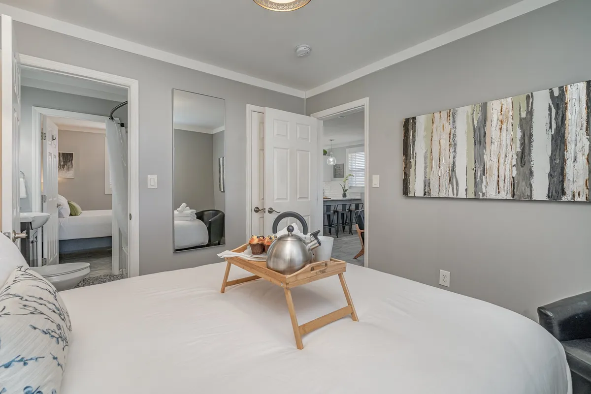 A serene bedroom with a queen-sized bed neatly made, a tray set for breakfast, tasteful wall art, and a mirror reflecting warm light. There are doorways visible that hint at easy access to other private areas, possibly the Jack-n-Jill bath or closet.