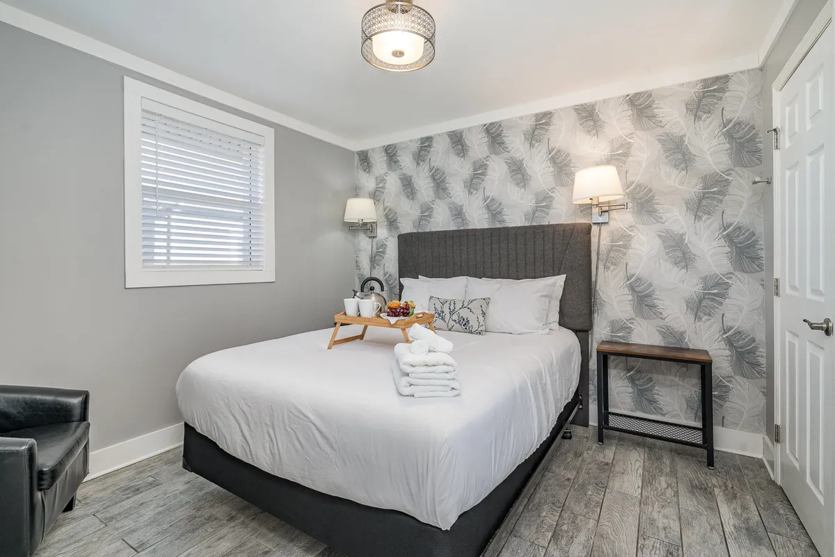 A cozy bedroom featuring a queen bed with decorative pillows, a breakfast tray with fruit and beverages, and a wall adorned with leaf-patterned wallpaper. The overall mood is comfortable and serene, ideal for relaxation and private retreats.